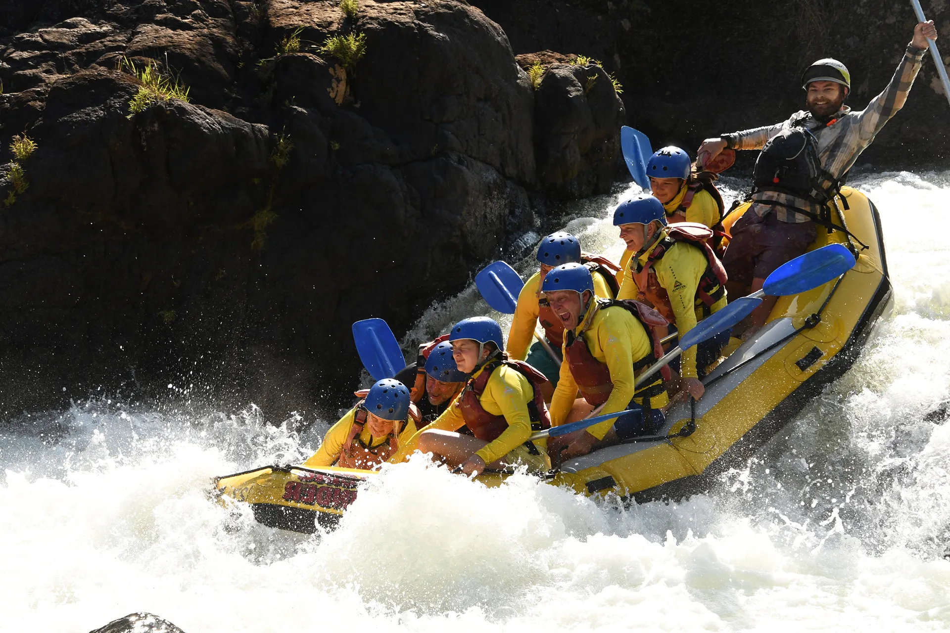 Group of people wearing helmets and life jackets navigating rough white water in a yellow raft.