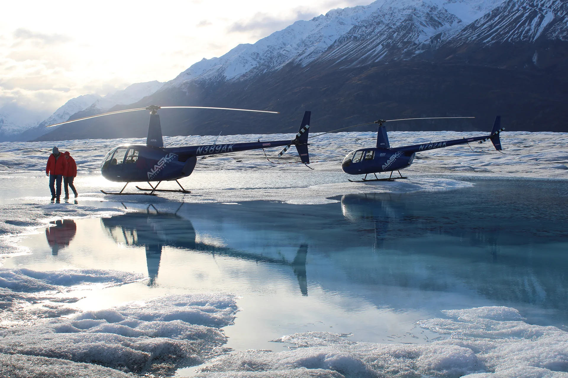 Two blue helicopters parked on a snowy glacier with two people in red jackets walking nearby and snow-capped mountains in the background.