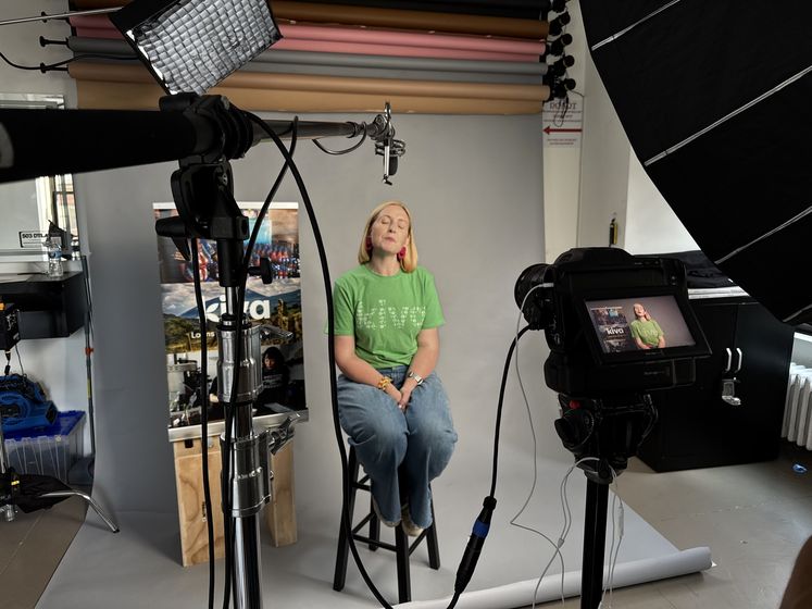 A staff member seated on a stool in a lit studio with the organization banner behind her, cameras and softboxes in the foreground