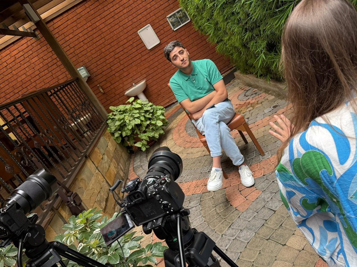 A Happy Productions interviewer talking with a young man in a brick courtyard, two cameras on tripods