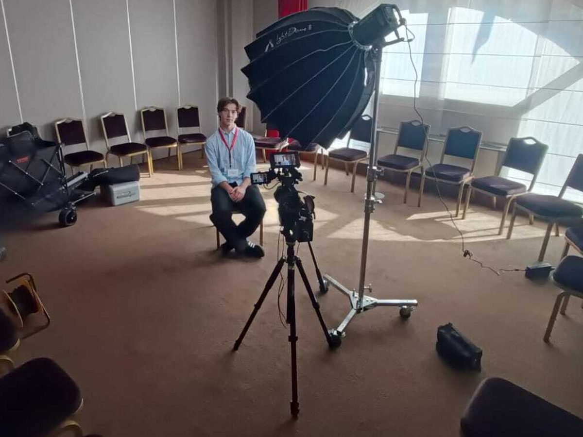 A young man wearing a lanyard seated under a large softbox, a camera set up in front of him