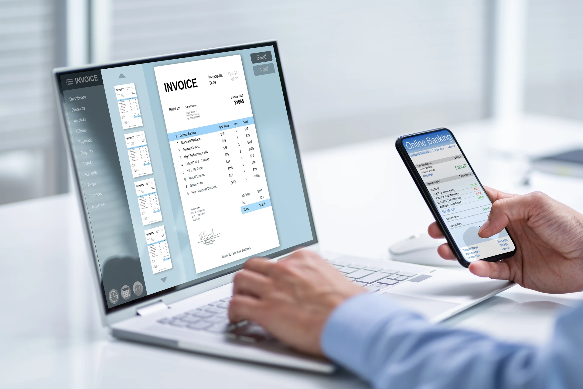 A person sits at a desk using a laptop that displays an invoice document, while holding a smartphone showing an online banking app