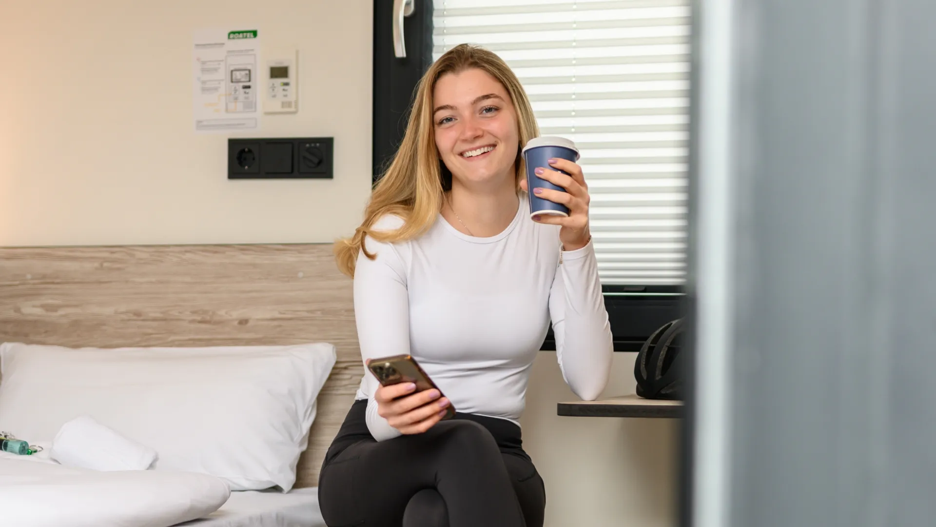 Junge Frau sitzt lächelnd mit Getränk und Smartphone in einem Zimmer.