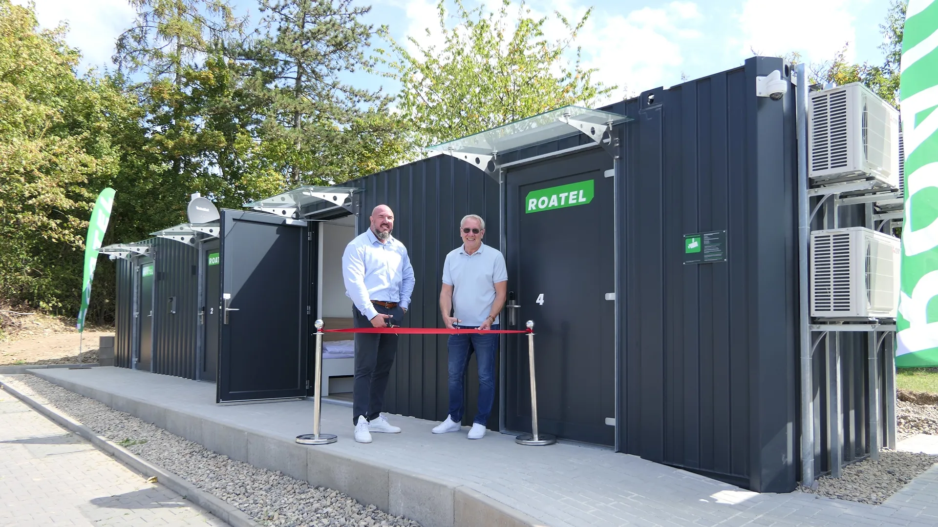 Zwei Männer stehen mit einer roten Eröffnungsschleife vor mehreren Roatel-Containern in Wolfhagen und halten Scheren für den offiziellen Ribbon-Cut.