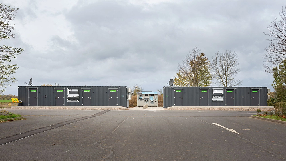 Roatel Anlage mit mehreren grauen Containern, die als Übernachtungsmöglichkeiten für Lkw-Fahrer dienen, auf einem Parkplatz mit Bäumen im Hintergrund.