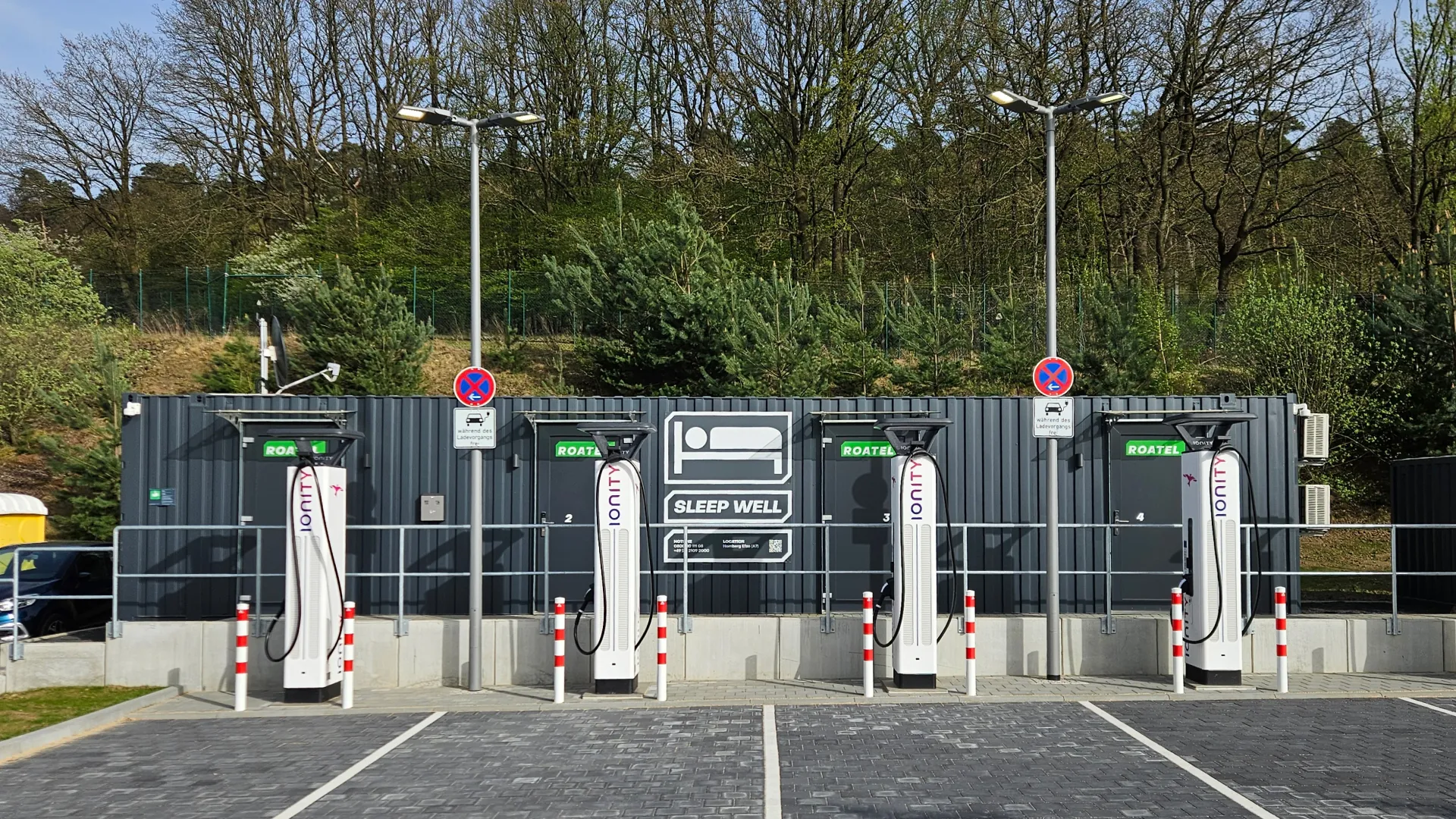 Vier IONITY-Ladestationen für Elektroautos vor einem Container mit dem Schriftzug 'Sleep Well' und Symbol eines Bettes, umgeben von Bäumen.