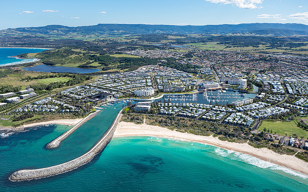 Aerial view of a coastal town with a marina, residential buildings, sandy beaches, and green hills in the background under a clear sky.