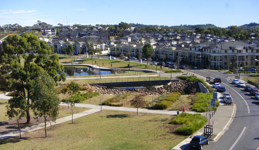 Residential neighborhood with modern houses, a small pond, green park area, and a curved road with parked cars.