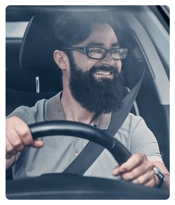 Homem branco de barba e usando óculos, está dirigindo, com as mãos no volante e olhando para direta.
