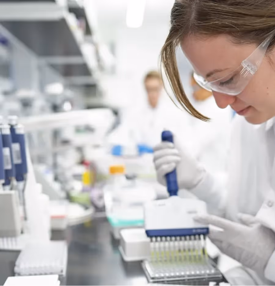 A scientist in a lab, wearing goggles, uses a pipette to transfer liquids, surrounded by lab equipment.