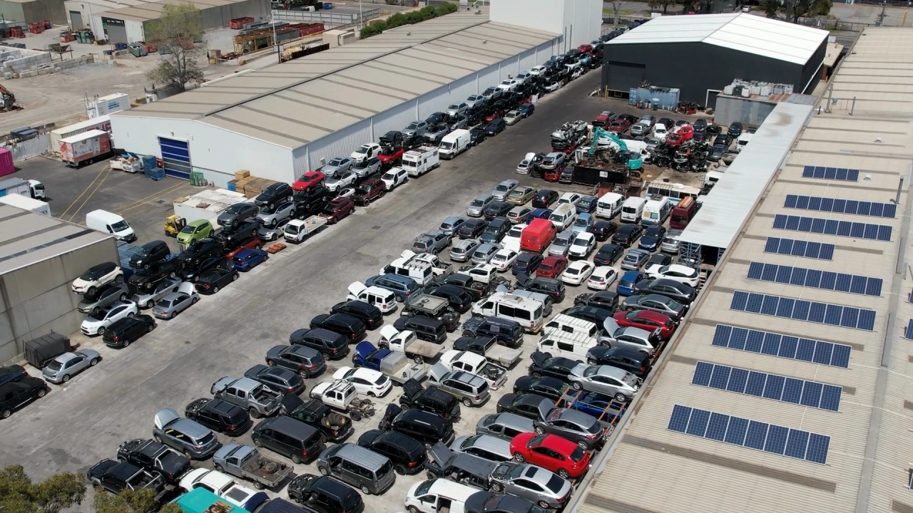 Aerial view of Wreckerland’s vehicle yard, showing rows of cars, vans, and trucks parked closely together, with some vehicles stacked. Surrounding industrial warehouses have solar panels installed on their roofs.