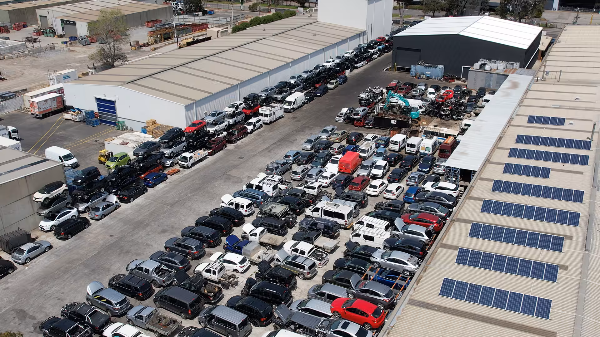 Aerial view of Wreckerland’s vehicle yard, showing rows of cars, vans, and trucks parked closely together, with some vehicles stacked. Surrounding industrial warehouses have solar panels installed on their roofs.