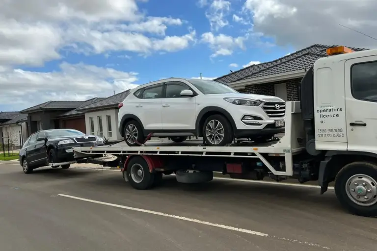 Tow truck transporting a white SUV on its flatbed with a black sedan partially lifted behind, parked on a suburban street with houses in the background.