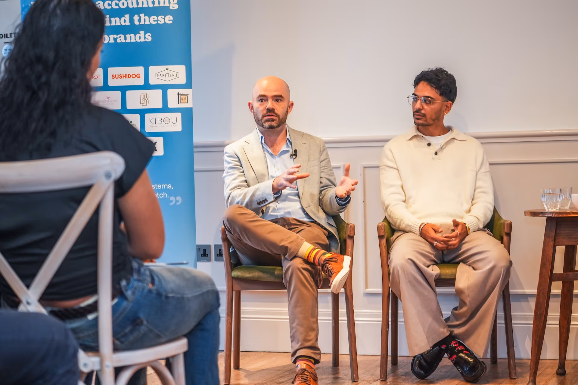 Two men seated and engaged in discussion during an indoor panel or meeting, with a person listening in the foreground.