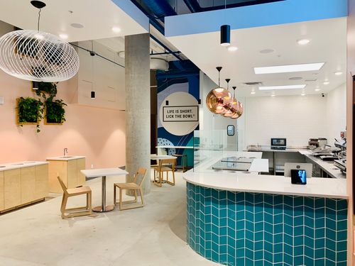 Interior of a contemporary café featuring pastel pink walls, teal tiled counters, wooden furniture, and modern decorative lighting.