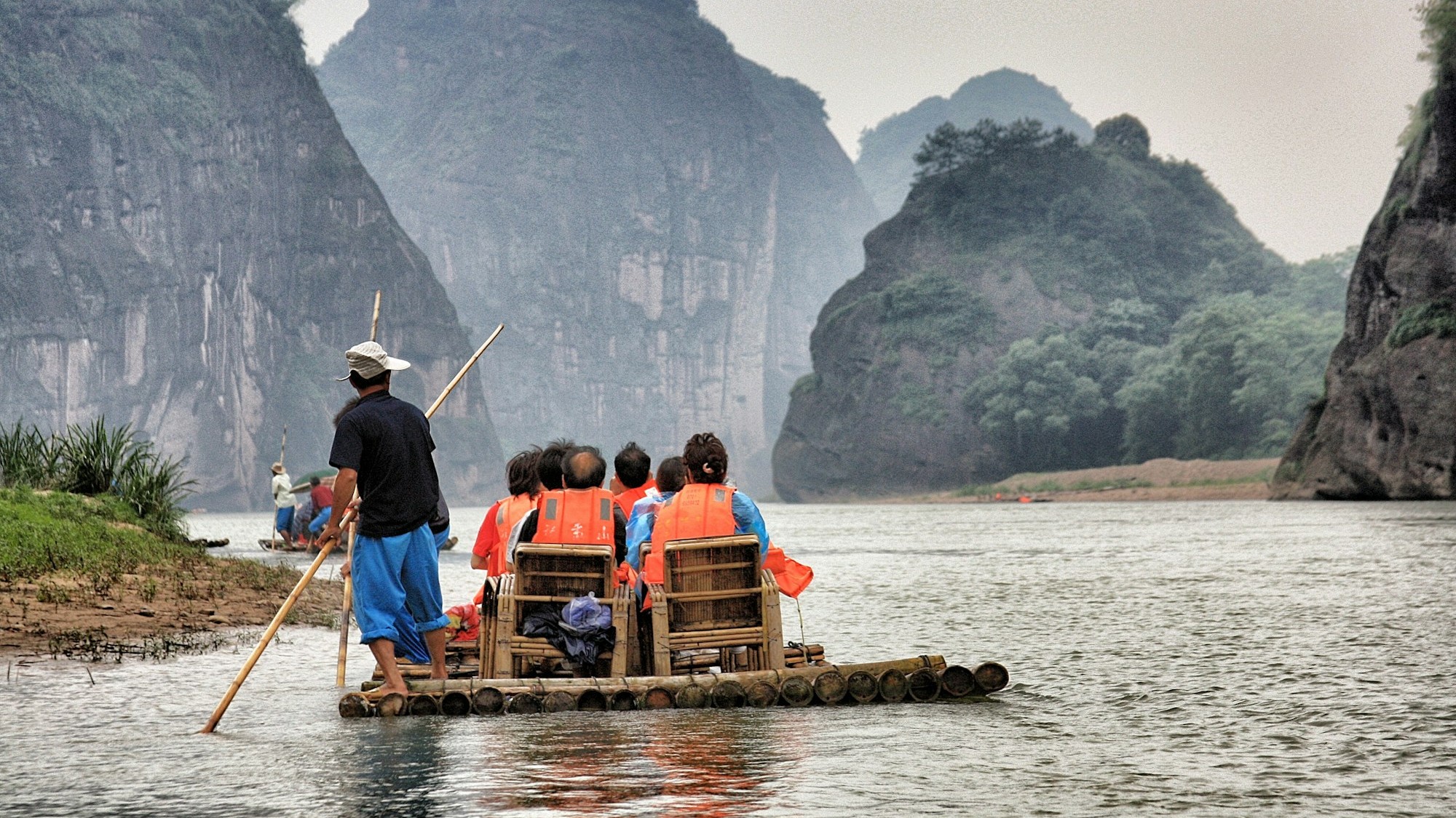 Guests in life jackets on a bamboo raft floating down a river between misty limestone cliffs