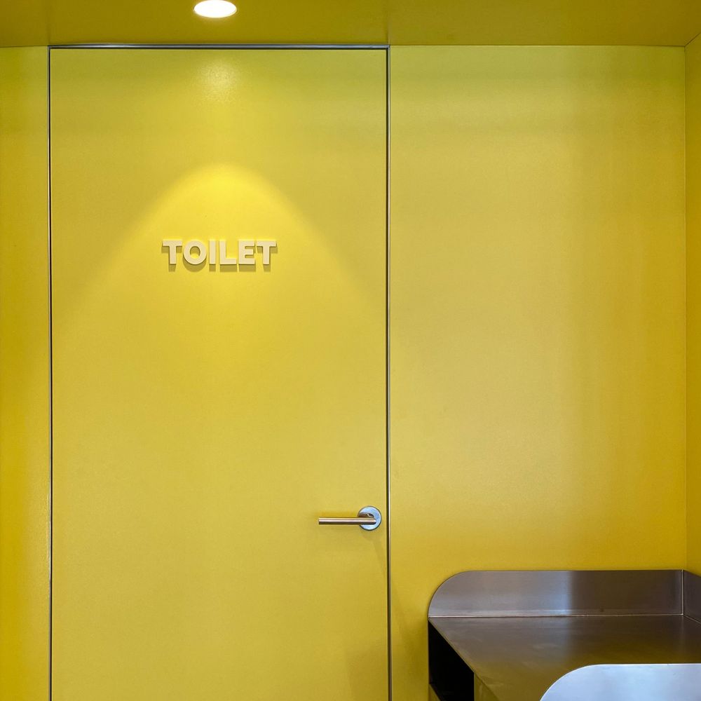 Gul dør med skiltet “TOILET” og dørhåndtag, oplyst af loftspot.