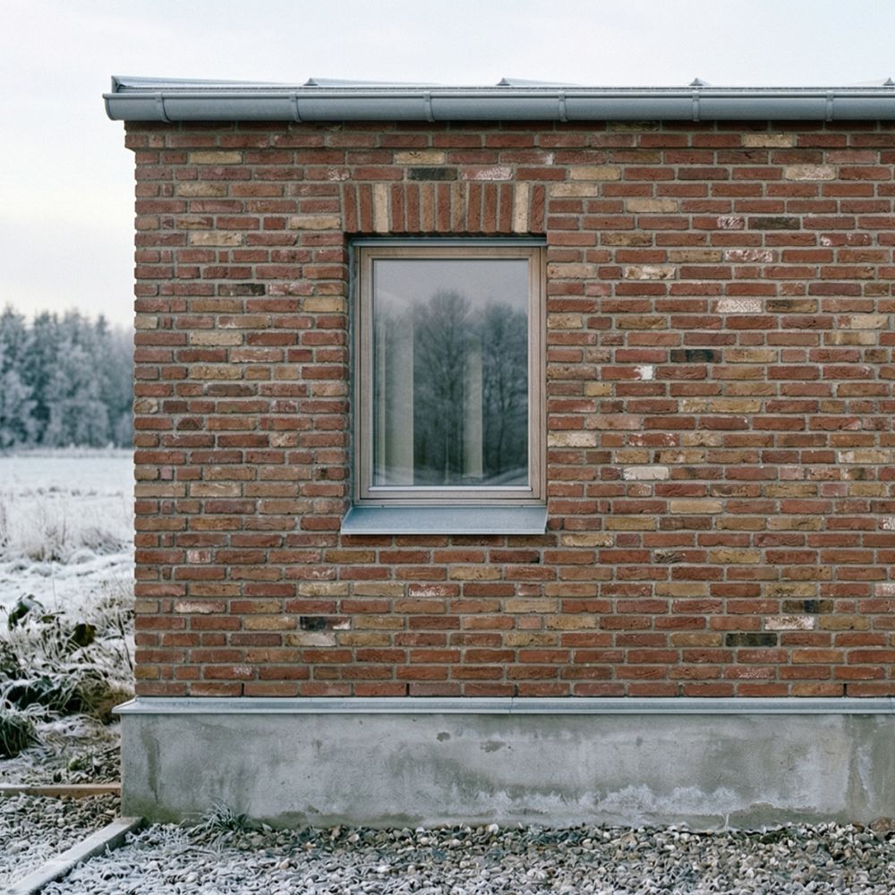 Murstensvæg med et enkelt vindue; vinterlandskab med frost og træer ses gennem ruden.
