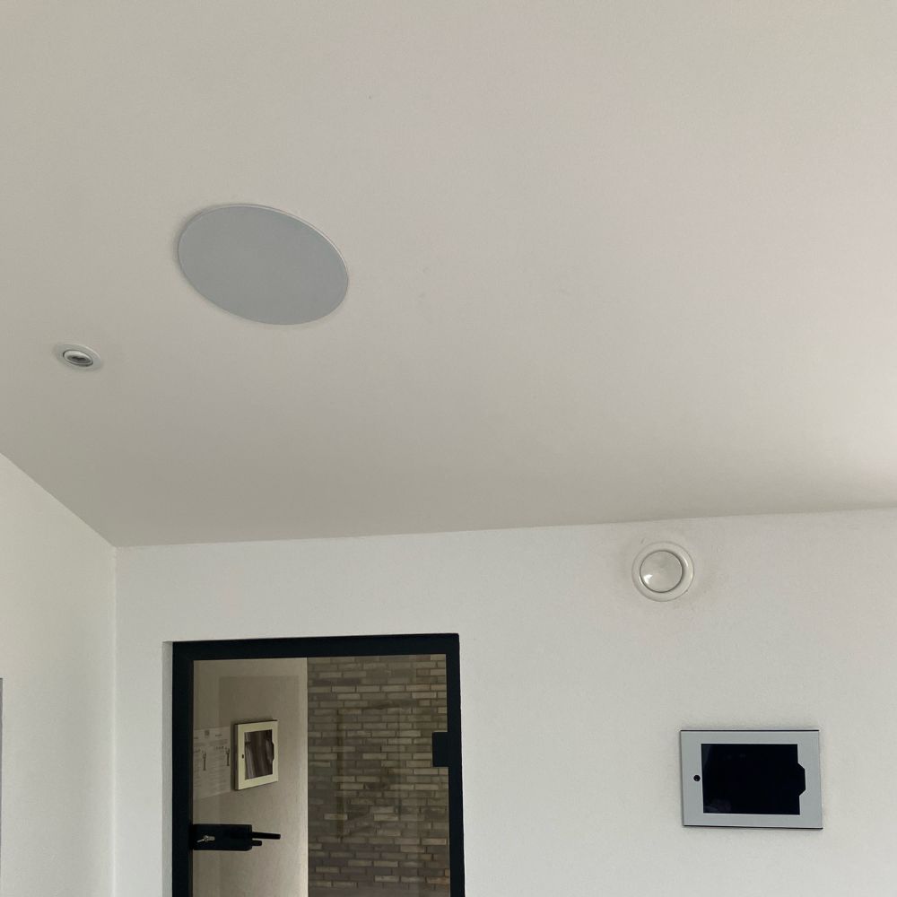 Hvidt loft og væg med indbygget højttaler, spot, ventilationsventil samt dør og vægpanel.