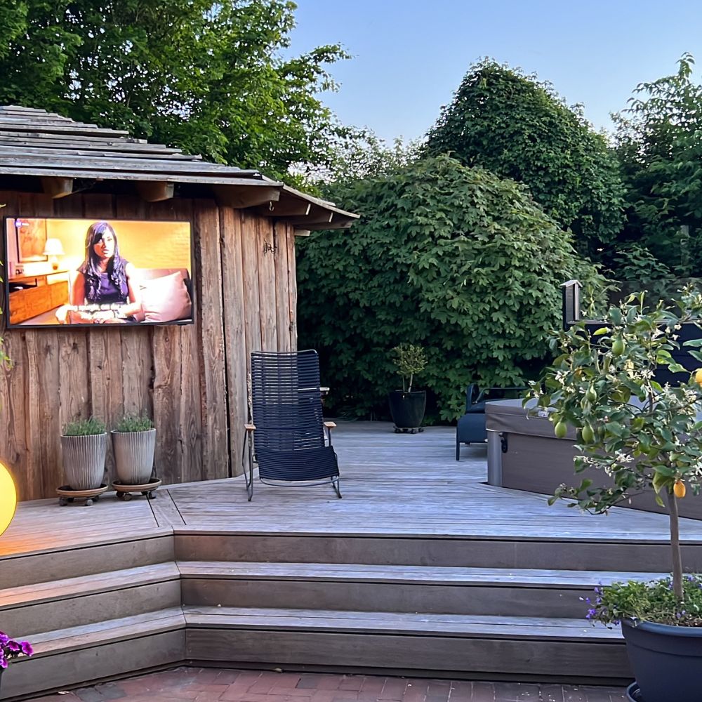 Træterrasse ved træskur med vægmonteret tv, stol og spa-badekar omgivet af grønne buske.