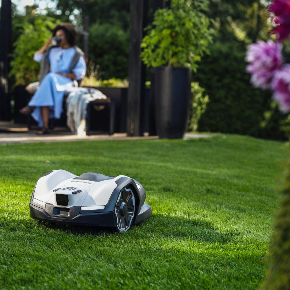 Robotplæneklipper kører på græsplæne i have, mens en person slapper af i baggrunden.