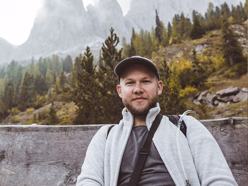 Man wearing a cap and light jacket sitting on a wooden bench with a forest and mountains in the background.
