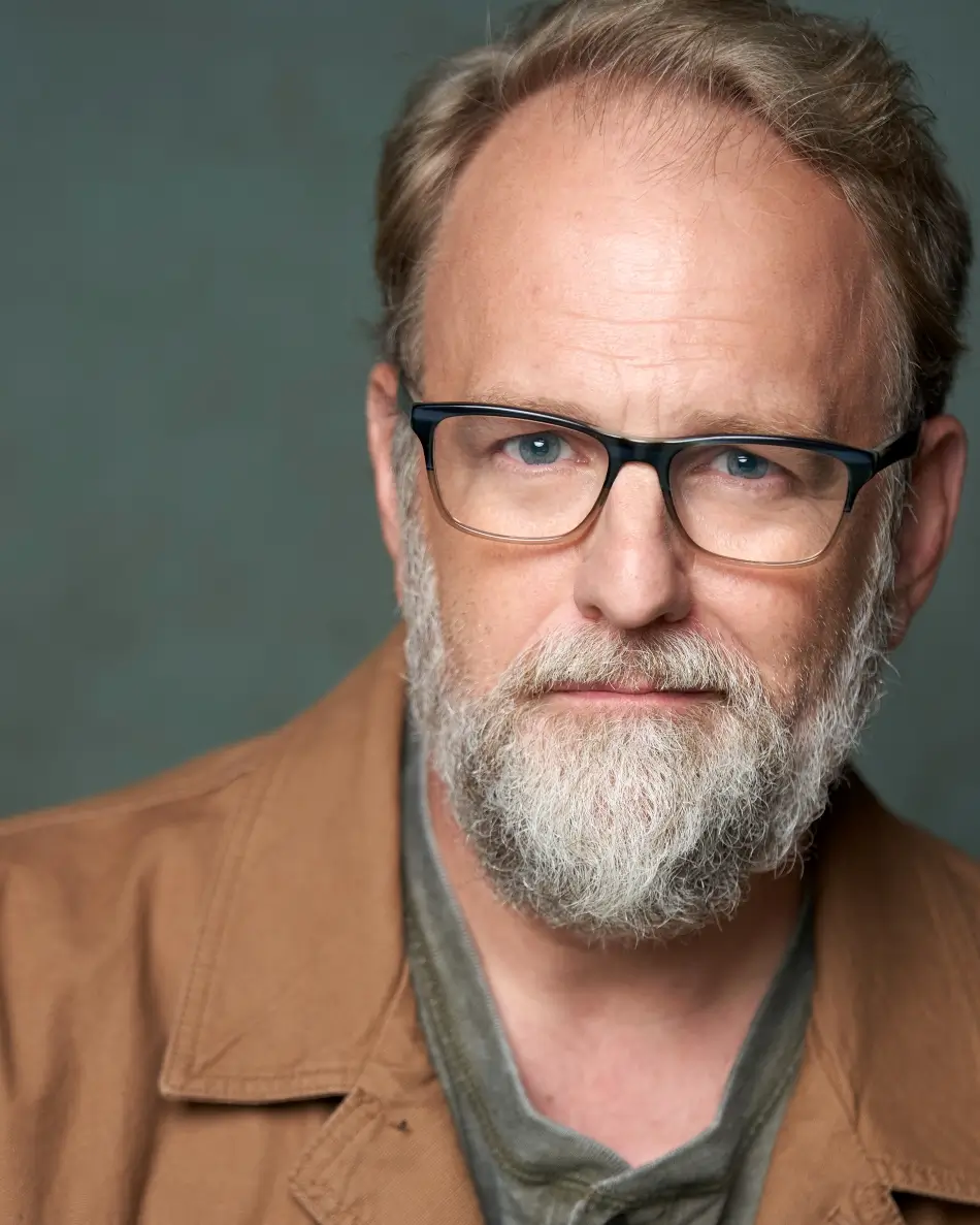 Atlanta men’s actor headshot — mature male with beard and glasses, serious expression in tan jacket and gray shirt against neutral background, authority and professional theatrical look.