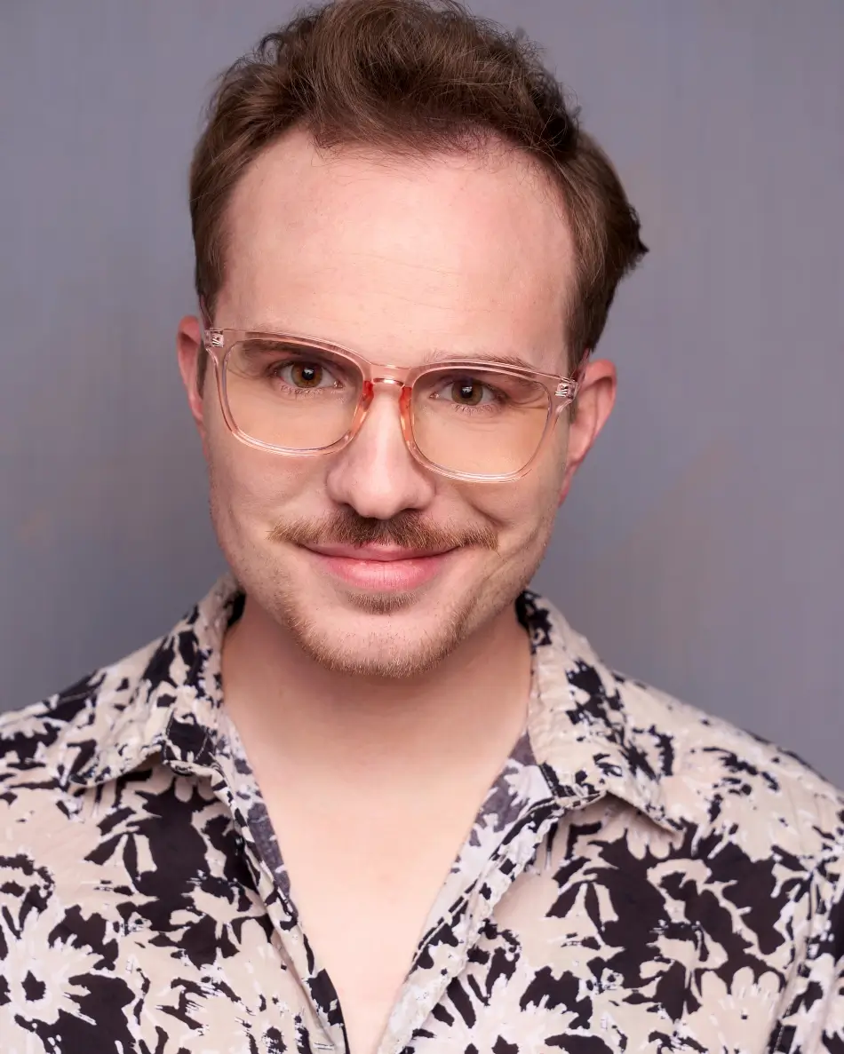 Atlanta men’s actor headshot — young male with translucent glasses and patterned shirt, playful smirk, quirky commercial character look against neutral background.