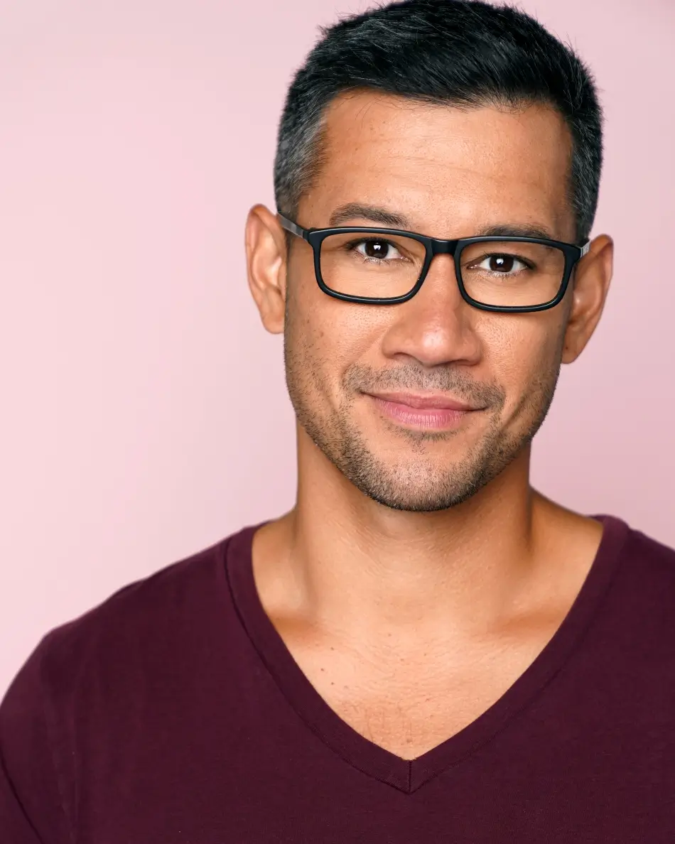 Atlanta men’s actor headshot —male with glasses and short dark hair, confident approachable smile in burgundy shirt against pink background, grounded commercial professional look.