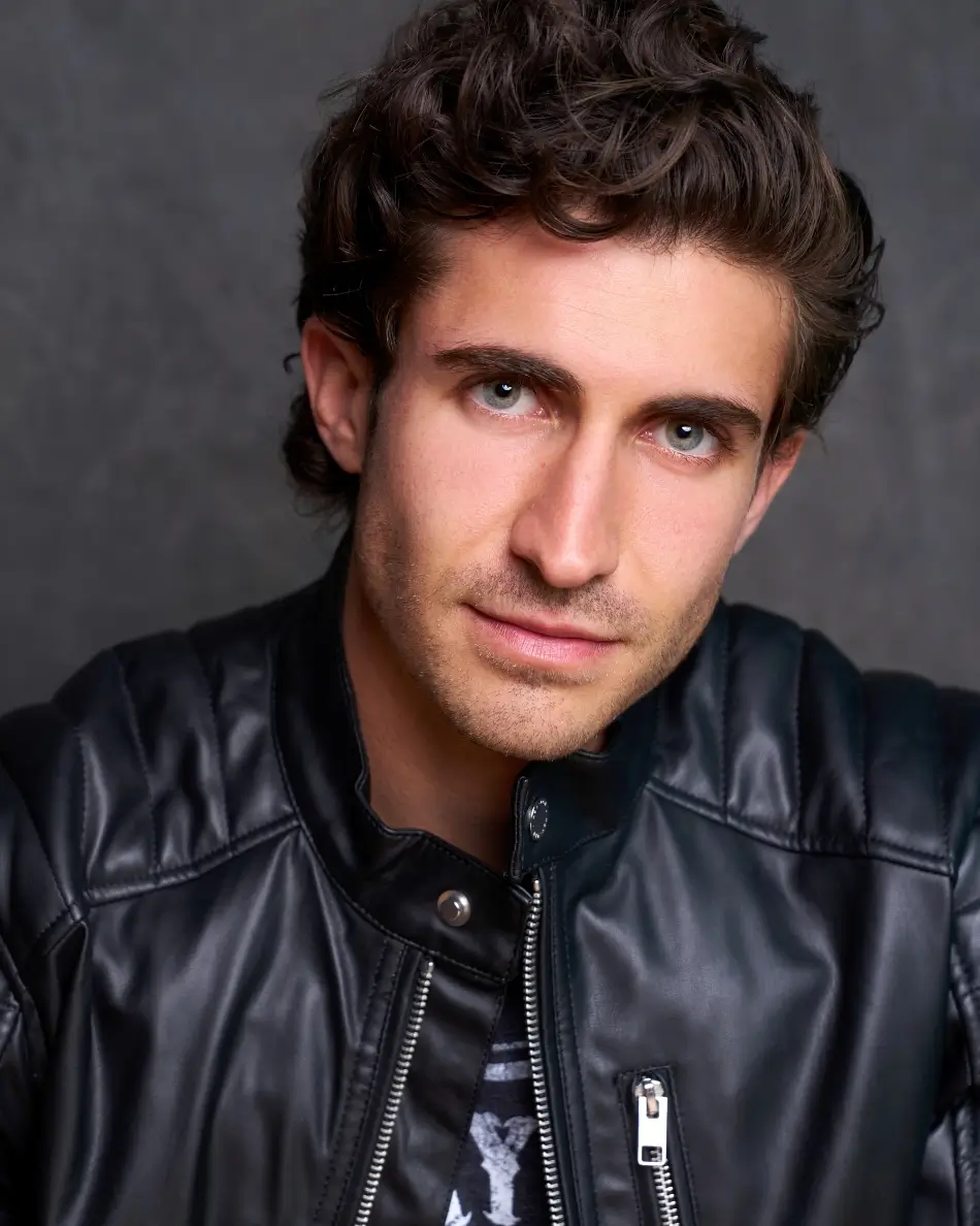 Atlanta men’s actor headshot — young male with curly dark hair and leather jacket, intense expression against dark background, edgy theatrical leading man look.