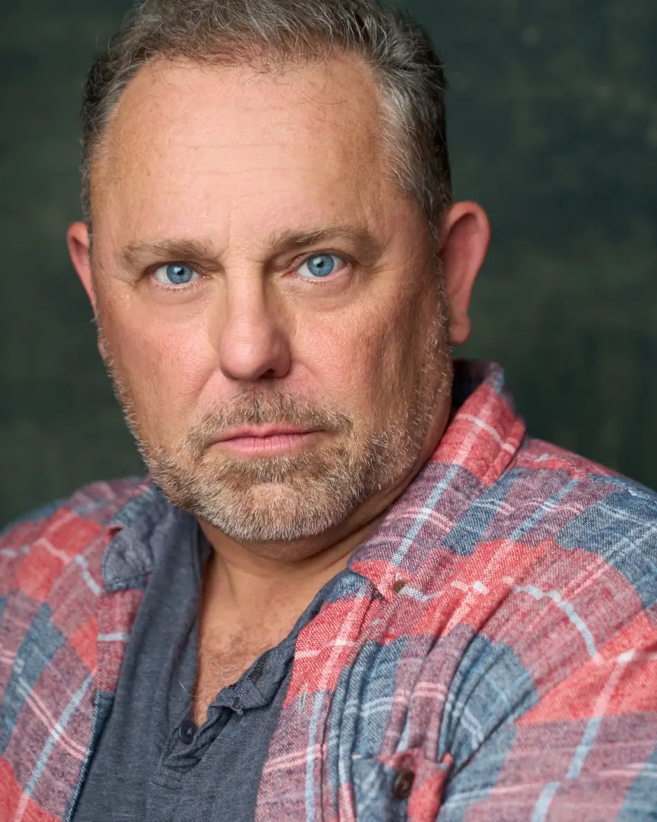 Atlanta men’s actor headshot — mature male with blue eyes, salt-and-pepper beard, serious expression in plaid shirt and henley, rugged theatrical authority look.