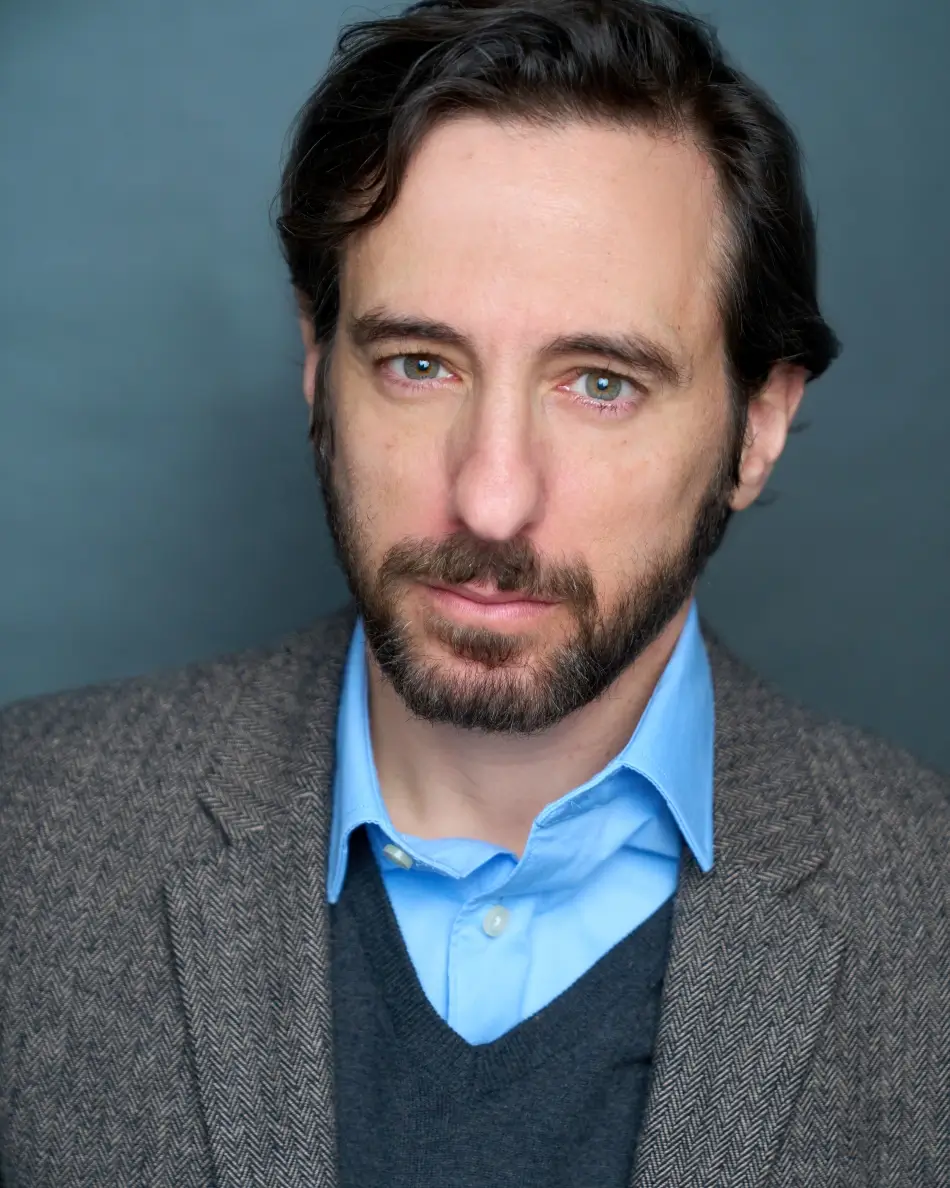 Atlanta men’s actor headshot — male actor with dark hair and beard, serious confident expression in blazer, sweater, and blue shirt, professional theatrical lawyer type look.