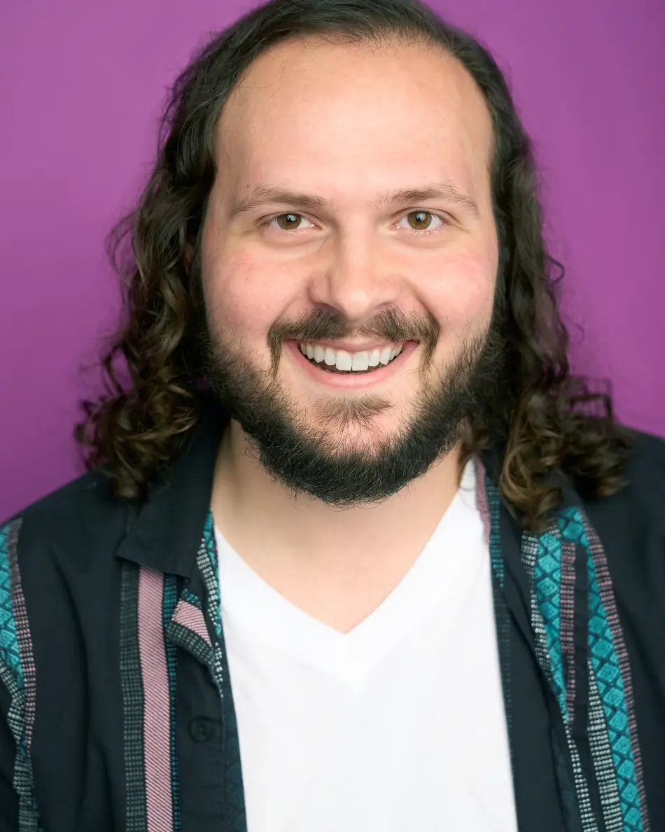 Atlanta men’s actor headshot — male actor with long dark hair and beard, bright smile in patterned shirt and white tee against purple background, commercial comedic and approachable look.