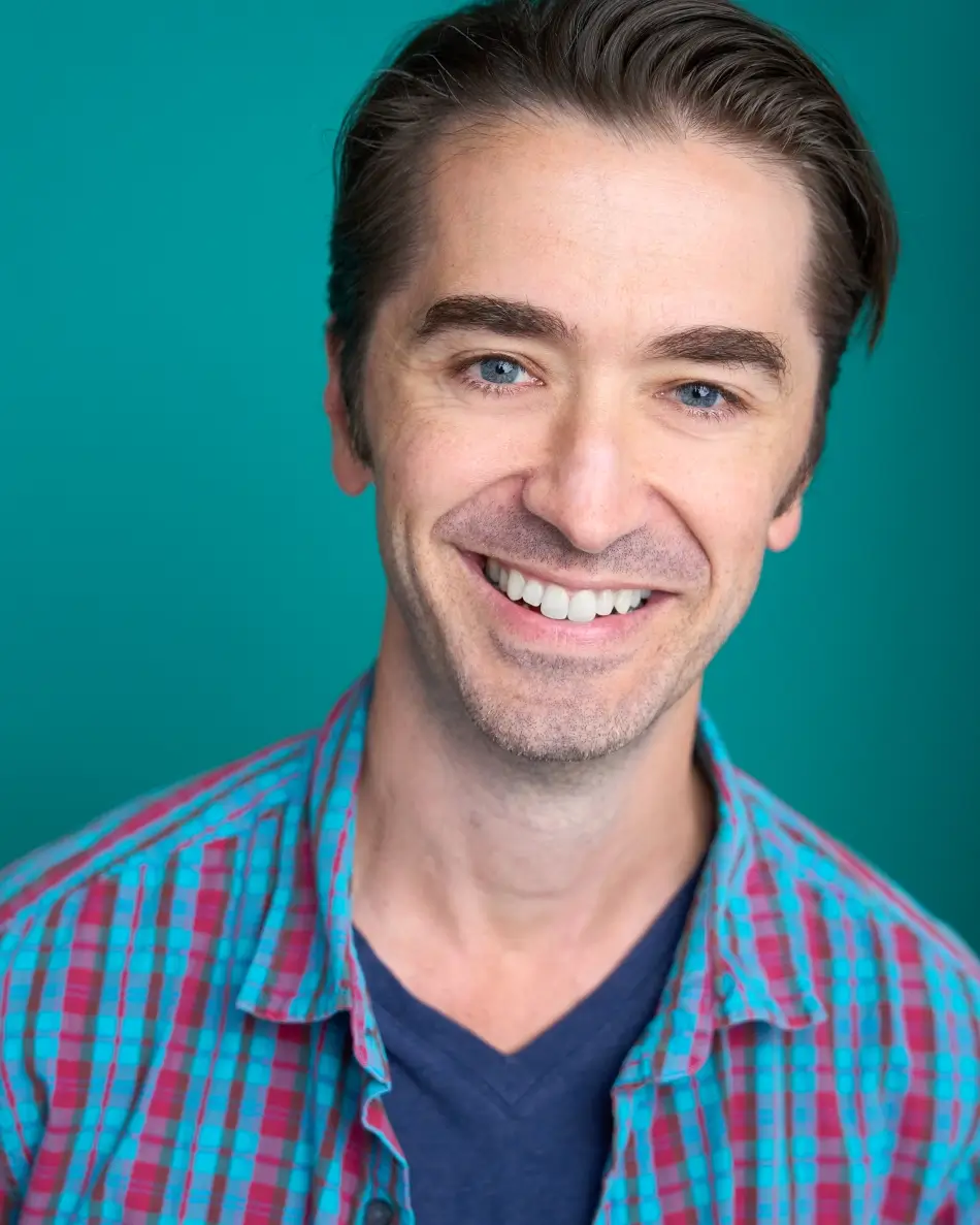 Atlanta men’s actor headshot — male actor with short dark hair, big smile in colorful plaid shirt and navy v-neck against teal background, bright friendly commercial lifestyle look.