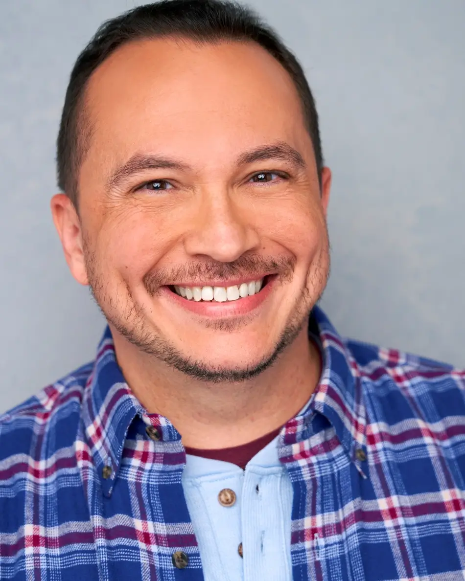 Atlanta men’s actor headshot — male actor with short dark hair and trimmed beard, big smile in plaid shirt against light neutral background, warm commercial everyman look.