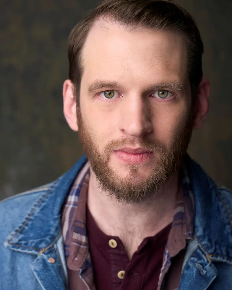 Atlanta men’s actor headshot — male actor with short dark hair and beard, calm but intense expression in denim jacket and plaid shirt against neutral background, rugged theatrical Americana look.