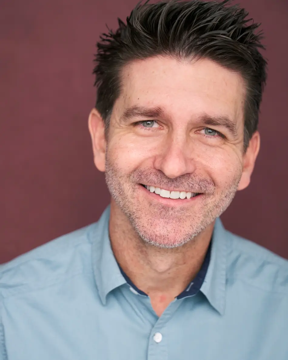Atlanta men’s actor headshot — male actor with short dark hair and beard, calm but intense expression in denim jacket and plaid shirt against neutral background, rugged theatrical Americana look.