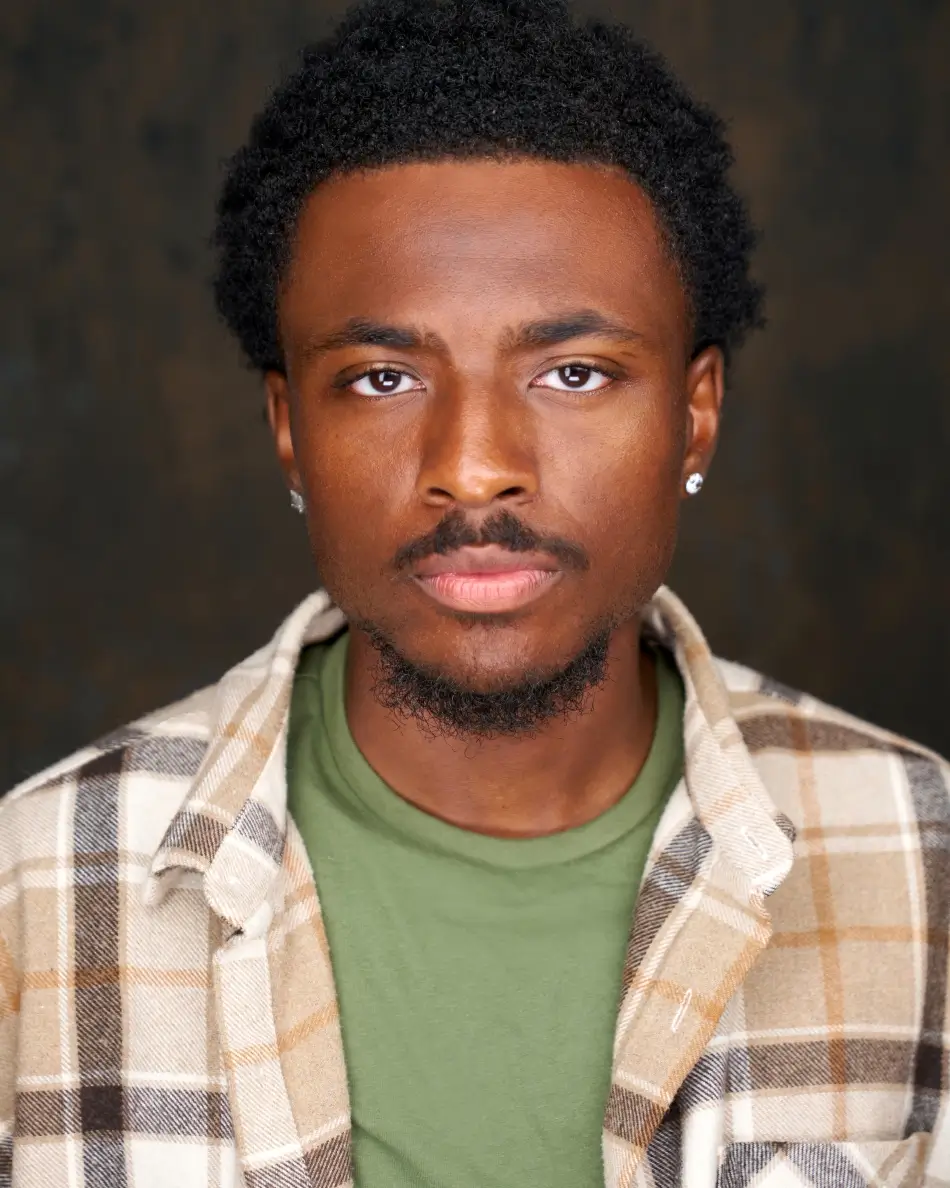 Atlanta men’s actor headshot — young male actor with short natural hair, serious expression in green shirt and plaid overshirt against dark background, grounded theatrical dramatic look.