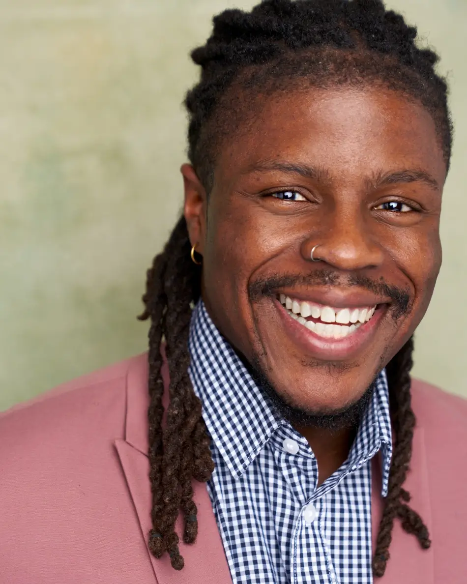 Atlanta men’s actor headshot — male actor with locs, nose ring, and big smile in pink blazer and gingham shirt against neutral background, charismatic commercial lifestyle look.