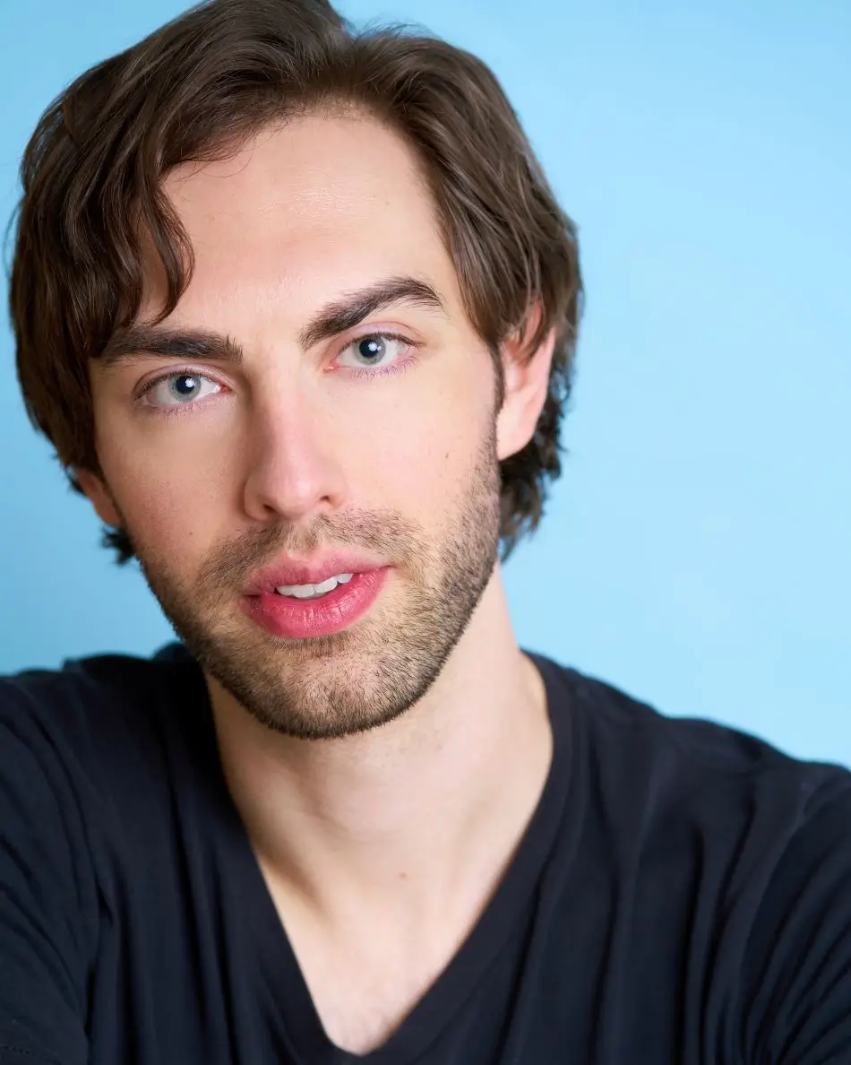 Atlanta men’s actor headshot — young male actor with brown hair and light stubble, serious but approachable expression in black shirt against light blue background, modern theatrical leading man look.
