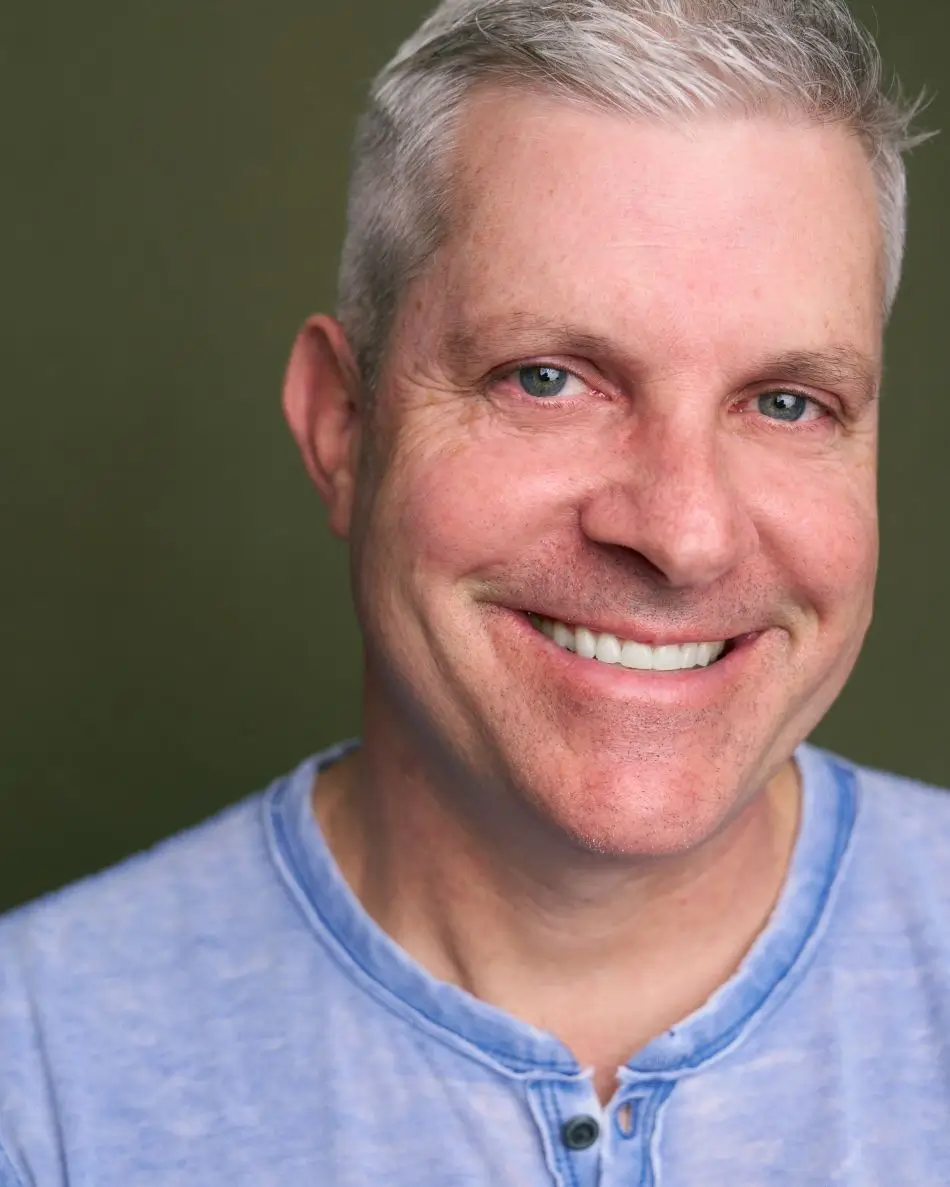 Atlanta men’s actor headshot — mature male with gray hair and blue eyes, warm smile in light-blue henley shirt against green background, friendly and trustworthy commercial look.