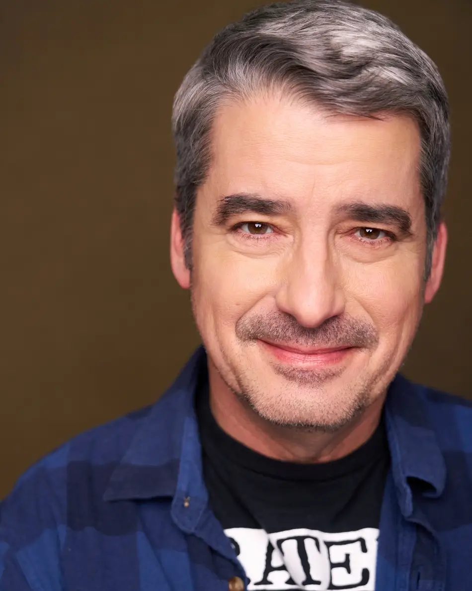 Atlanta men’s actor headshot — mature male with salt-and-pepper hair, subtle confident smile in plaid shirt and graphic tee against neutral background, grounded theatrical character look.