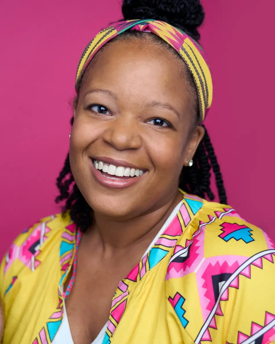Atlanta commercial actor headshot — female actor with braids and colorful headband, bright toothy smile in vibrant yellow patterned top against pink background.