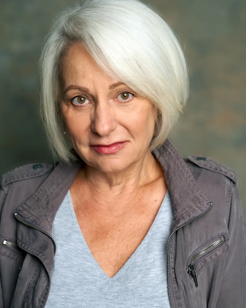 Atlanta theatrical actor headshot — mature female actor with white hair, serious expression in gray utility jacket and v-neck top, dramatic and authority role type.