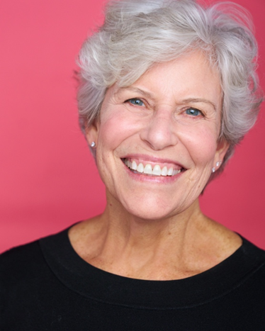Atlanta commercial actor headshot — mature female actor with short white hair, bright toothy smile in black top against pink background, friendly and welcoming look.