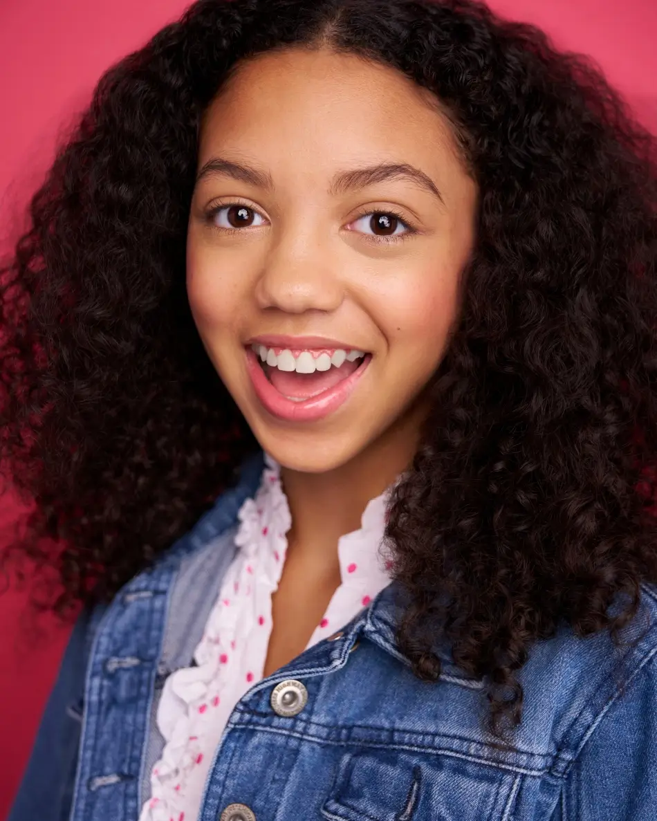 Atlanta tween actor headshot — tween girl with curly hair, expressive smile in denim jacket and ruffled shirt against pink background, commercial headshot for tween roles.
