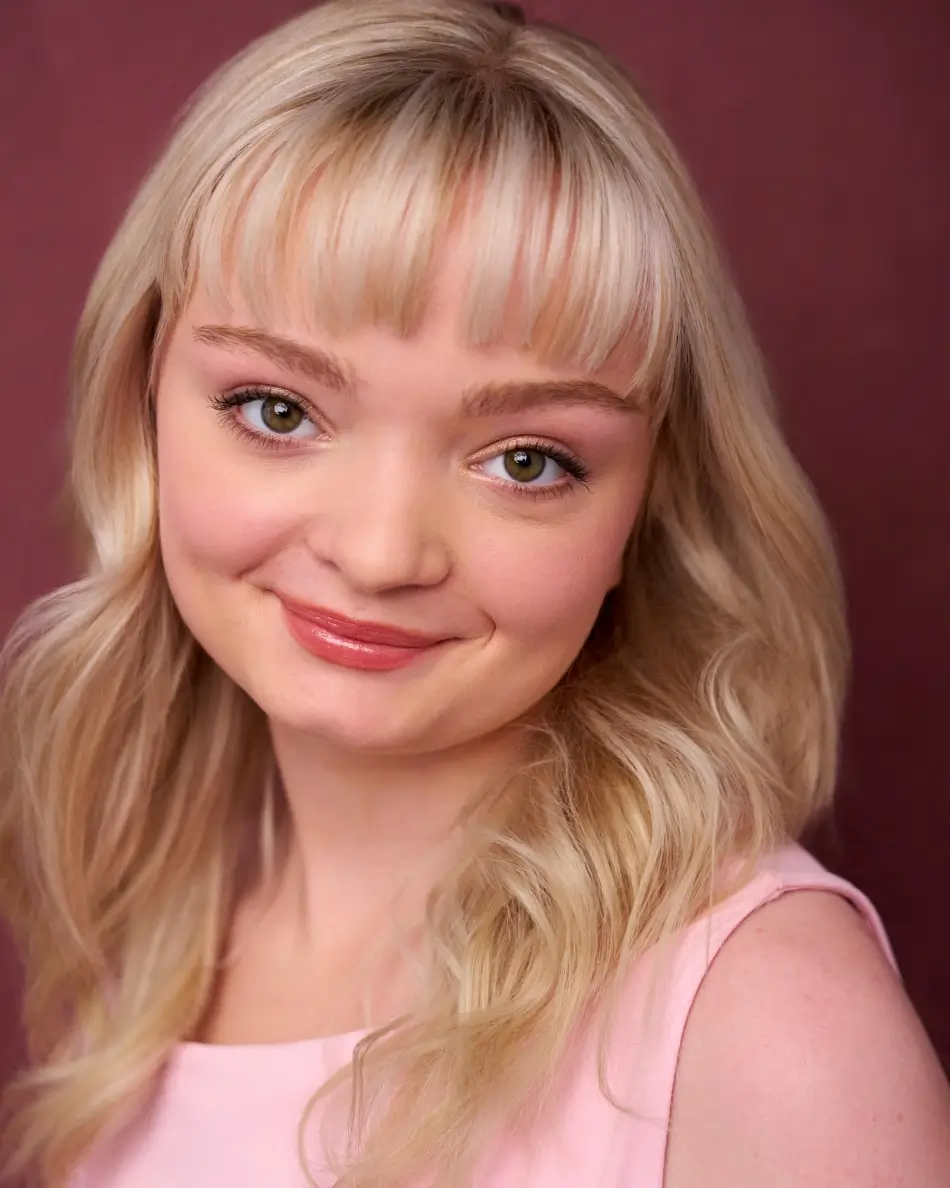 Atlanta teen actor headshot — teen girl with long blonde hair and bangs, soft smile in pink dress against maroon background, polished warm commercial teen look.