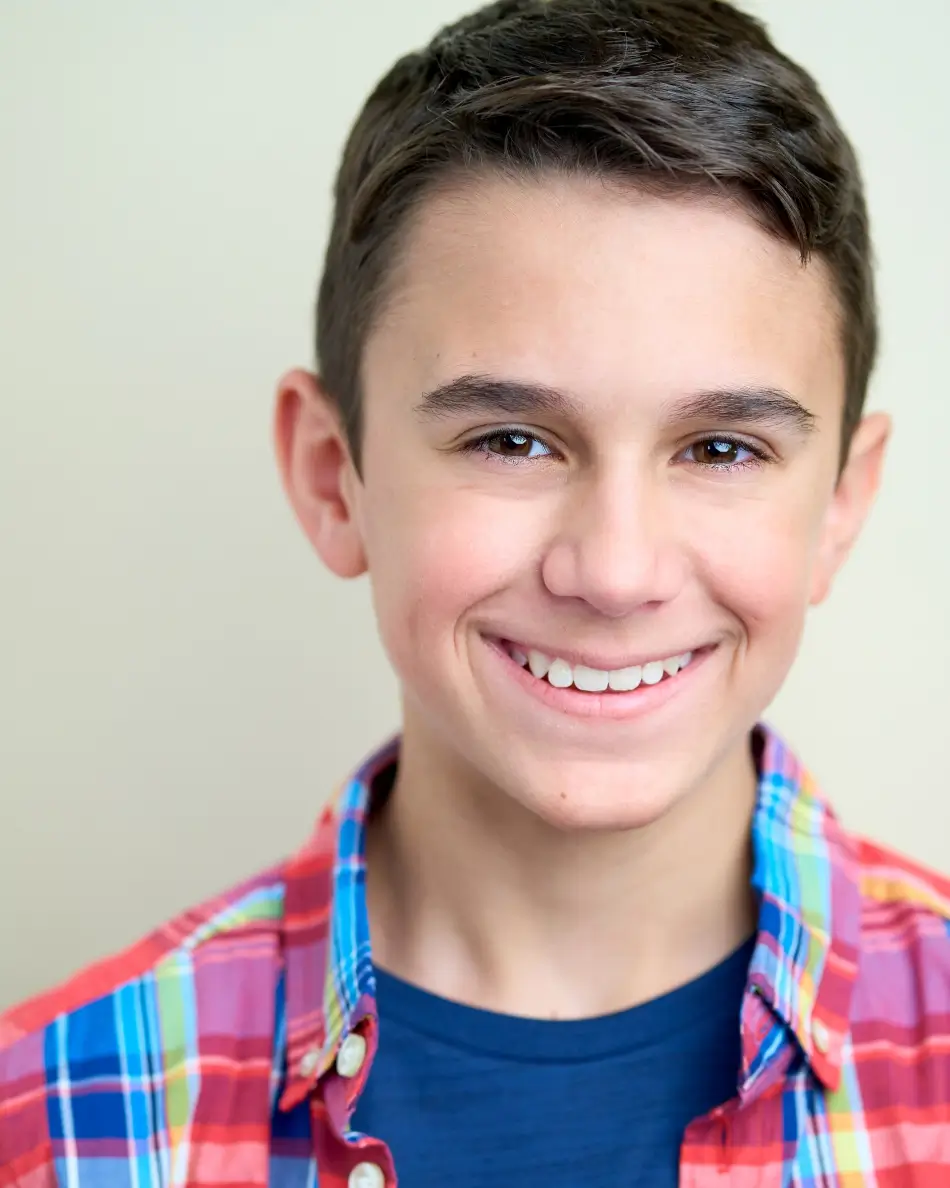 Atlanta teen actor headshot — teen boy with short dark hair, bright approachable smile in colorful plaid shirt and navy t-shirt against light background, commercial-friendly teen look.