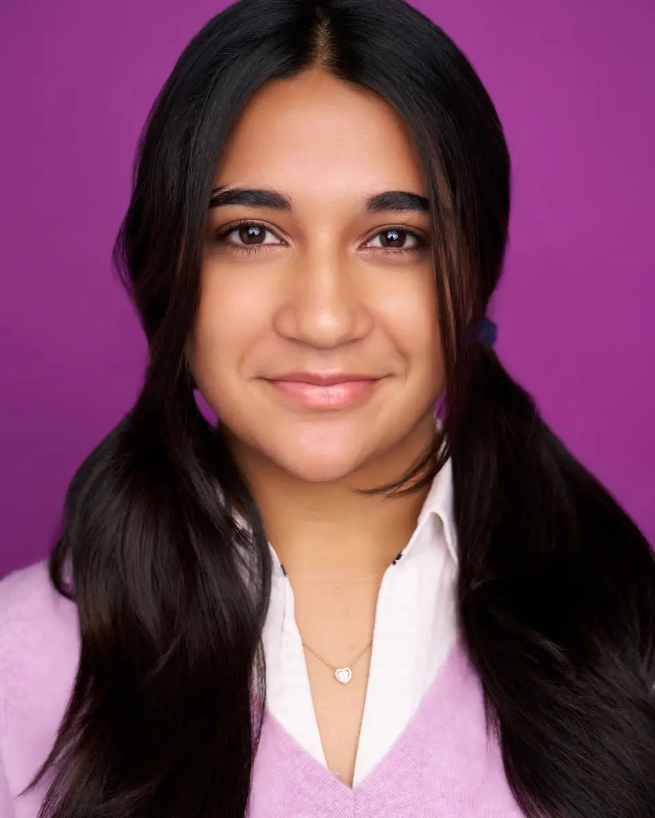 Atlanta teen actor headshot — teen girl with long dark hair in pigtails, subtle smile in purple sweater and collared shirt against purple background, polished approachable commercial teen look.