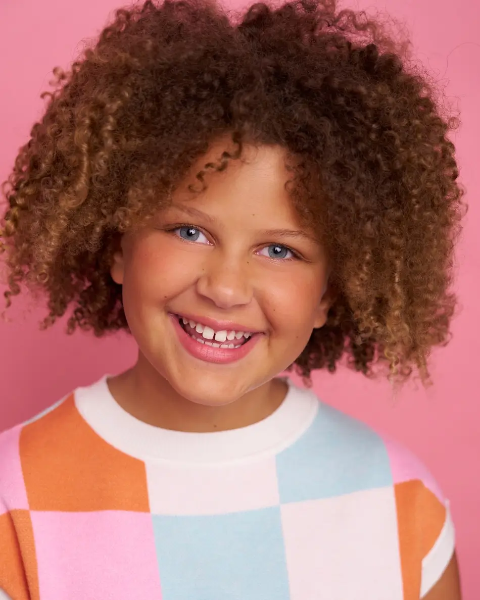 Atlanta kids actor headshot — young child with curly hair and big smile in colorful pastel checkerboard top against pink background, bright cheerful commercial kids look.